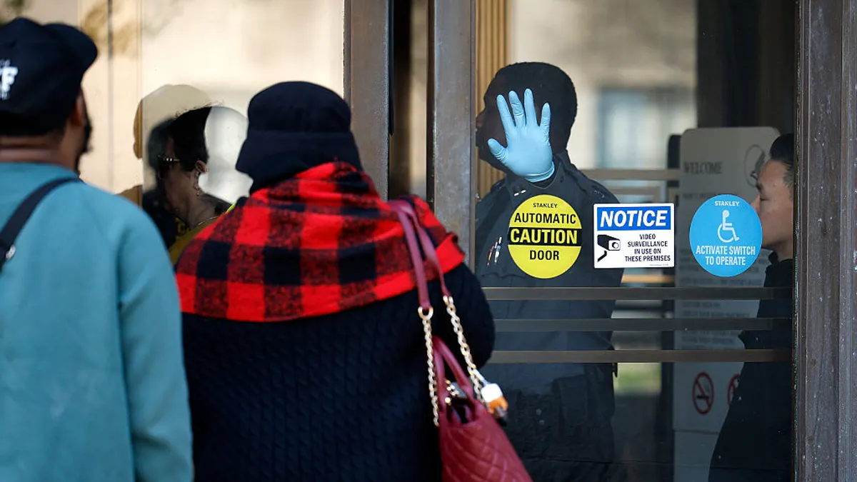 People wait in line outside of a glass door. A person in uniform holds a gloved hand to the door.