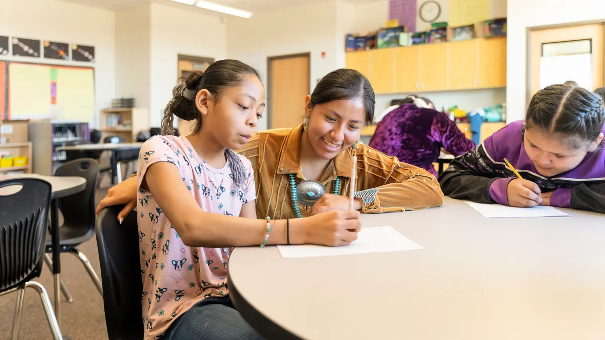 An adult is with two students at a desk in a classroom. The students are writing and reading.