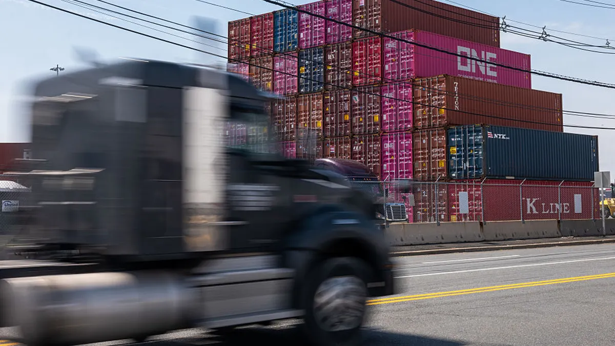The cab of a semi truck appears blurry while driving past a stack of shipping containers in the background.