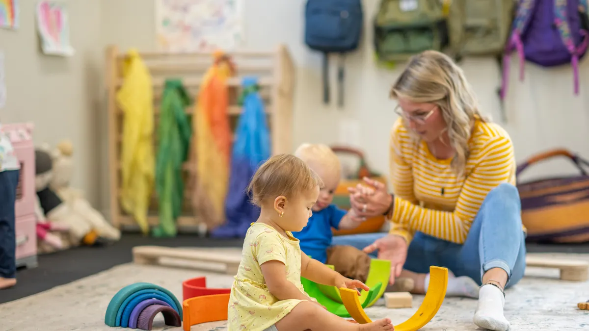 An adult is sitting on a floor with two toddlers who are sitting on the floor. The adult is guiding one of the child's hands while the other child plays with blocks on the floor.