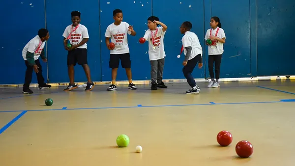 A handful of younger students are in a school gym playing bocce with balls.