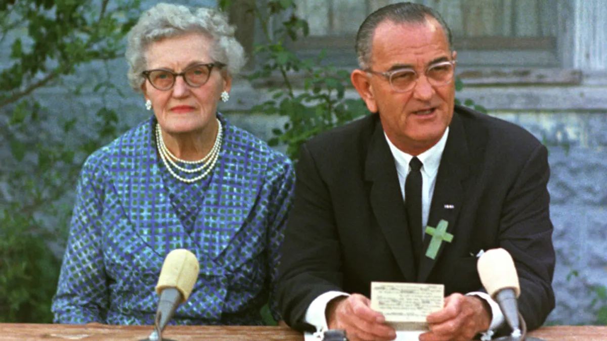 Two people dressed in business attire are seated at a desk outside with a building behind them.